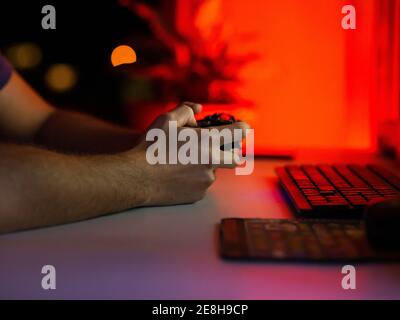 Rognez un joueur masculin méconnaissable à l'aide d'une manette de jeu et en jouant à un jeu vidéo assis à une table avec ordinateur dans une pièce sombre Banque D'Images