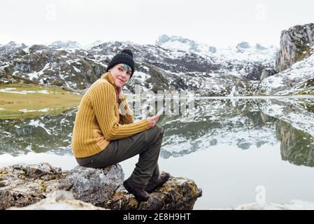 Vue latérale sur tout le corps jeune femme heureuse portant un pull chaud et un casque à l'aide d'un téléphone portable et en regardant loin lorsque vous êtes assis sur la rive du lac de poney pointu surroun Banque D'Images