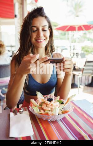 Jeune femme photographiant sa salade avec un smartphone tout en étant assise dans un restaurant Banque D'Images