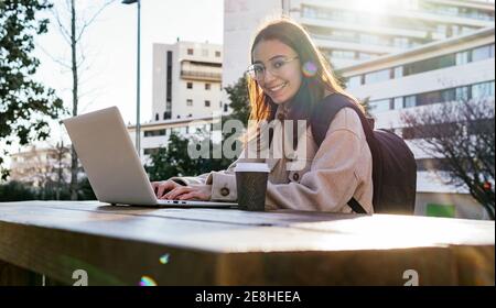 Bonne femme souriante étudiante en manteau chaud et lunettes avec sac à dos pour travailler à la maison sur netbook tout en étant assis table avec tapis à emporter Banque D'Images