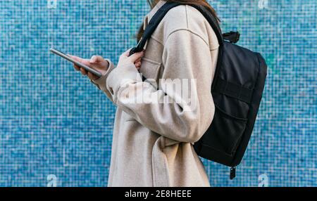 Vue latérale d'une étudiante inreconnaissable dans une tenue décontractée avec sac à dos posé contre un mur carrelé bleu Banque D'Images