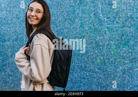 Vue latérale petit élève femme en tenue décontractée avec sac à dos se tenir contre un mur carrelé bleu et regarder loin avec le sourire Banque D'Images