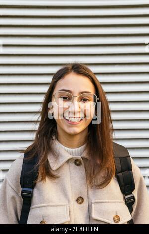 Vue avant petit élève femme en tenue décontractée avec sac à dos debout dans la rue et regardant la caméra avec le sourire Banque D'Images