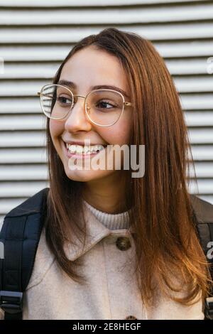 Vue avant petit élève femme en tenue décontractée avec sac à dos se tenir dans la rue et regarder loin avec le sourire Banque D'Images