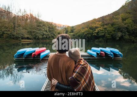 Vue arrière jeune couple aimant en tenue chaude embrassant chacun d'autres tendinement en se tenant sur un quai en bois de lac contre la végétation luxuriante abondantes collines boisées dans le re Banque D'Images