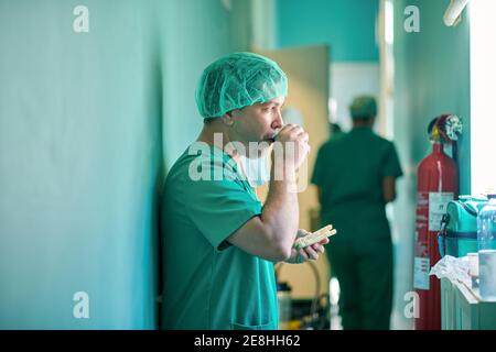 Vue latérale d'un jeune médecin masculin concentré en uniforme médical et de boucher boire du café à emporter et manger un sandwich après la chirurgie Banque D'Images