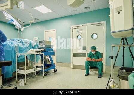 Anonyme jeune homme médecin en uniforme médical vert et masque envoyez des messages sur votre téléphone mobile lorsque vous êtes assis dans la salle d'opération après chirurgie Banque D'Images
