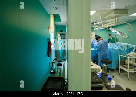 Vue latérale du praticien anonyme de sexe masculin dans les exfoliations smartphone dans le couloir tandis que le groupe de médecins effectuant la chirurgie dans salle d'opération Banque D'Images