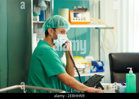 Vue latérale du jeune homme concentré médic dans les gommages et masque médical parlant au téléphone et regardant loin à l'hôpital quartier Banque D'Images
