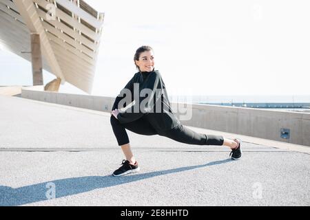 Pleine longueur déterminée jeune Sportswoman en activewear étirant les jambes tout en se réchauffant sur la route dans la banlieue Banque D'Images