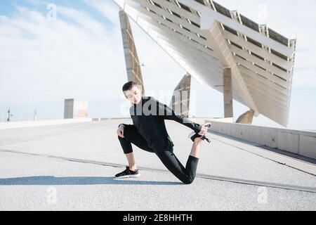 Pleine longueur déterminée jeune Sportswoman en activewear étirant les jambes tout en se réchauffant sur la route dans la banlieue Banque D'Images