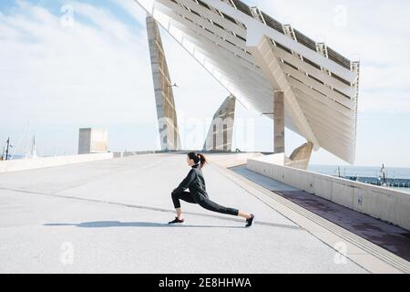 Pleine longueur déterminée jeune Sportswoman en activewear étirant les jambes tout en se réchauffant sur la route dans la banlieue Banque D'Images
