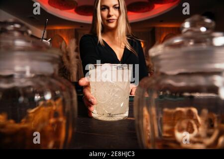 Contenu jeune femme barman avec de longs cheveux blonds servant froid cocktail alcoolisé dans un restaurant élégant qui regarde l'appareil photo Banque D'Images