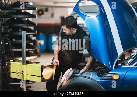 Jeune homme d'entretien barbu concentré répare la voiture contemporaine contre Bright lumière et vue sur l'extérieur dans l'atelier Banque D'Images
