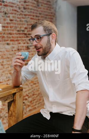 Barista masculin en chemise blanche et avec une petite tasse de expresso assis au comptoir en bois dans le café et vue loin Banque D'Images