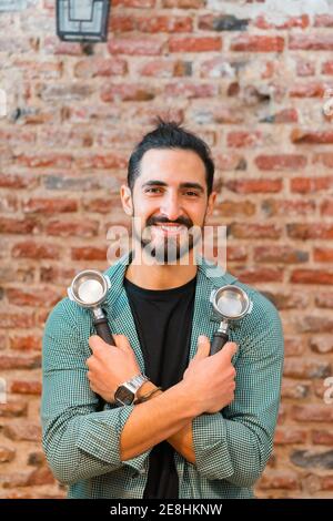 Un barista homme joyeux avec des portafilters regardant l'appareil photo en arrière-plan de mur de briques dans le café style loft Banque D'Images