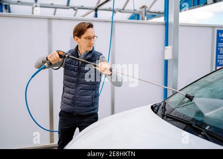 Jeune homme concentré dans une tenue décontractée et lunettes de vue lave-glace de voiture avec pistolet à eau dans la rue Banque D'Images
