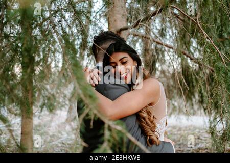 Vue latérale des jeunes mariés avec de longs cheveux robe blanche souriant et embrassant groom avec les yeux fermés pendant se tenant ensemble près de l'arbre de conifères Banque D'Images