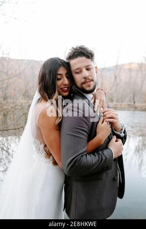 Vue latérale des jeunes mariés avec de longs cheveux robe blanche souriant et embrassant groom avec les yeux fermés pendant se tenant ensemble près de l'arbre de conifères Banque D'Images