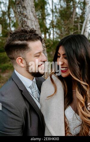 Vue latérale d'un jeune gai à barbe en costume riant et une élégante mariée ethnique en robe blanche tout en étant debout ensemble sur pont en bois n Banque D'Images