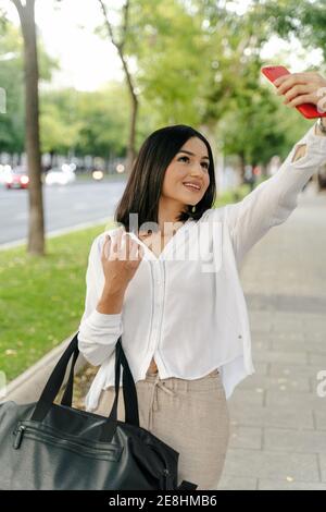 Charmante femme enchantée se tenant dans la rue de la ville et se prenant soi-même tourné sur un smartphone pendant une promenade le week-end Banque D'Images