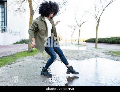 Vue latérale de la jeune femme afro-américaine expressive millénaire tenue tendance plongeant le pied dans l'eau de la flaque tout en restant debout rue par beau temps Banque D'Images