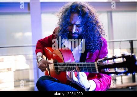 Homme ethnique concentré avec de longs cheveux jouant de la guitare acoustique pendant répéter la chanson sur scène à la lumière des projecteurs Banque D'Images