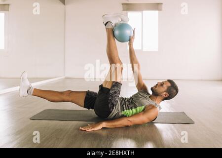 Vue latérale d'un jeune sportif musclé déterminé en activité Exercice de Crunch de jambe avec Medicine ball tout en étant allongé sur le tapis pendant l'entraînement en lumière Banque D'Images