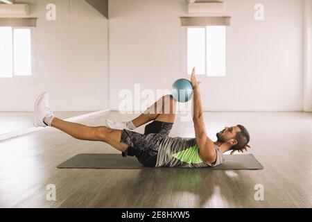 Vue latérale d'un jeune sportif musclé déterminé en activité Exercice de Crunch de jambe avec Medicine ball tout en étant allongé sur le tapis pendant l'entraînement en lumière Banque D'Images