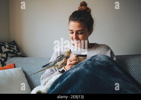 Bonne jeune femme dans un chandail chaud souriant et buvant chaud café tout en vous relaxant sur un canapé avec un adorable oiseau de cafard main Banque D'Images