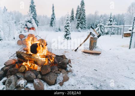 Théière placée sur un feu de camp sur un champ enneigé près des conifères contre le ciel nuageux au coucher du soleil en hiver Banque D'Images