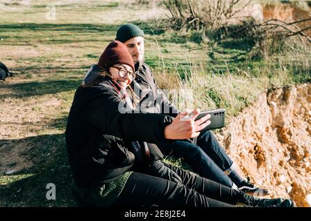 Vue latérale d'un couple de touristes heureux en train de prendre des vêtements d'extérieur autoportrait sur téléphone portable tout en restant sur le support contre le ravin rétro-éclairé Banque D'Images