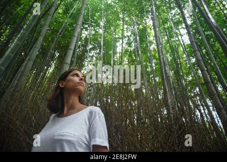 Jeune femme caucasienne attrayante regarde et admire de grands arbres dans la forêt de bambous d'Arashiyama à Kyoto, au Japon Banque D'Images