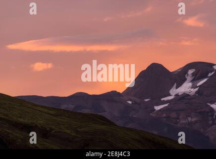 Vue à couper le souffle sur les majestueuses montagnes accidentées avec de la neige sur les pistes près de la vallée herbeuse vallonnée sous le pittoresque ciel rose nocturne dedans Islande Banque D'Images