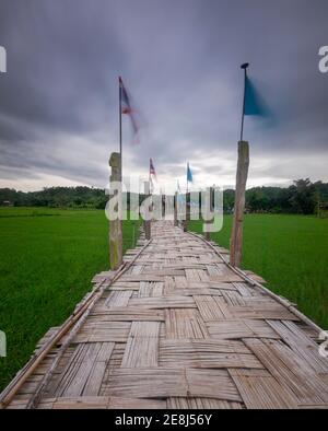 Long pont en bambou su Tong PAE avec différents drapeaux sur piliers en bois traversant le champ de riz contre le ciel nuageux Thaïlande Banque D'Images