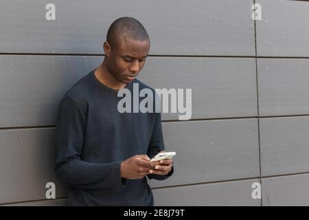 Un homme afro-américain attentif fait défiler un téléphone mobile moderne tout en étant debout contre le bâtiment gris Banque D'Images