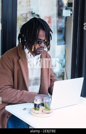 Un homme noir hipster sérieusement ciblé dans des lunettes de soleil avec des dreadlocks assis à la table avec ordinateur portable tout en travaillant sur le projet à distance dans restaurant extérieur Banque D'Images