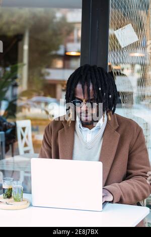 Un homme noir hipster sérieusement ciblé dans des lunettes de soleil avec des dreadlocks assis à la table avec ordinateur portable tout en travaillant sur le projet à distance dans restaurant extérieur Banque D'Images