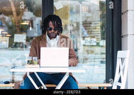 Un homme noir hipster sérieusement ciblé dans des lunettes de soleil avec des dreadlocks assis à la table avec ordinateur portable tout en travaillant sur le projet à distance dans restaurant extérieur Banque D'Images