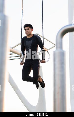 Corps entier bas angle de jeune sportif asiatique en noir vêtements d'entraînement accrochés à des anneaux de gymnastique lors d'exercices sur le sport masse Banque D'Images