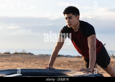 Jeune homme asiatique déterminé dans les vêtements de sport effectuant l'exercice de poussée contre le pneu du tracteur pendant les entraînements intenses en extérieur Banque D'Images