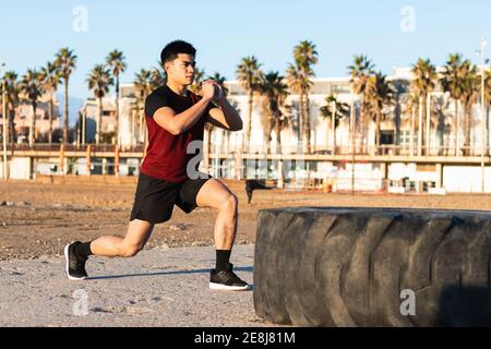 Vue latérale complète du corps d'un jeune homme asiatique déterminé vêtements de sport effectuant des fentes tout en vous réchauffant et en vous préparant à faire de l'exercice avec pneu de voiture sur la broche Banque D'Images