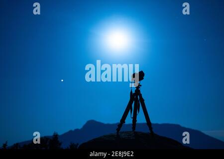 Appareil photo professionnel avec lanière sur trépied contre la silhouette de montagne sous un ciel coloré avec le soleil au crépuscule Banque D'Images