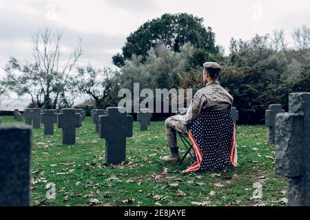 Vue arrière du corps du soldat en uniforme assis chaise avec drapeau américain tout en pleurant la mort des guerriers à cimetière Banque D'Images