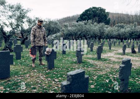 Corps complet de soldat en uniforme debout avec le drapeau américain tout en pleurant la mort de guerriers au cimetière Banque D'Images