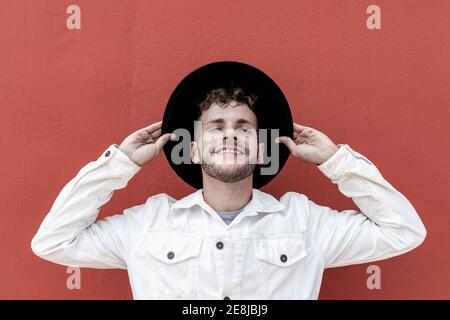 Joyeux jeune homme millénaire dans des vêtements élégants ajuster chapeau et souriant avec les yeux fermés tout en se reposant sur la rue contre le rouge mur Banque D'Images