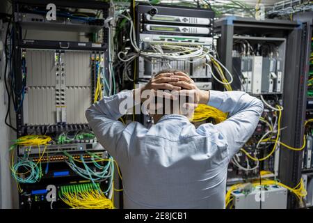 Vue arrière d'un technicien mâle méconnaissable qui attrane la tête problème dans le centre de données avec les racks de serveurs Banque D'Images