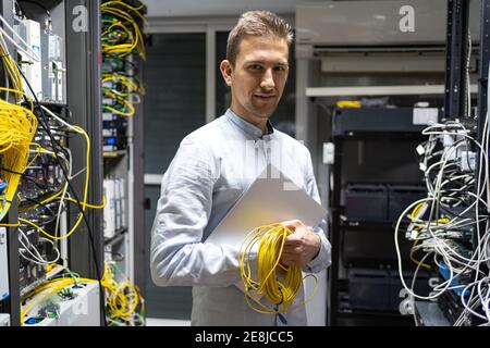 Homme positif dans un casque sans fil debout avec un netbook et des câbles dans la salle des serveurs Banque D'Images