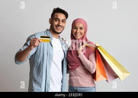 Banque fiable. Couple musulman joyeux posant avec une carte de crédit et des sacs de shopping Banque D'Images