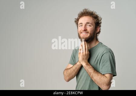 Mendiant pour aider à mettre les mains ensemble comme dans la prière beau cheveux bouclés souriant homme regardant l'appareil-photo étain besoin habillé en t-shirt olive isolé sur blanc Banque D'Images